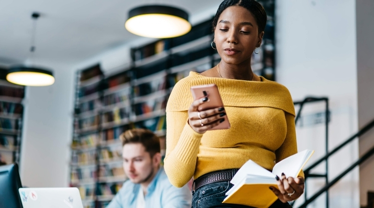 An adult woman holding a hand while flipping through a book in a library. There is another person seated behind her working as well