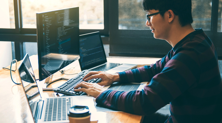 An adult man working on his laptop with two laptops next to him at his desk