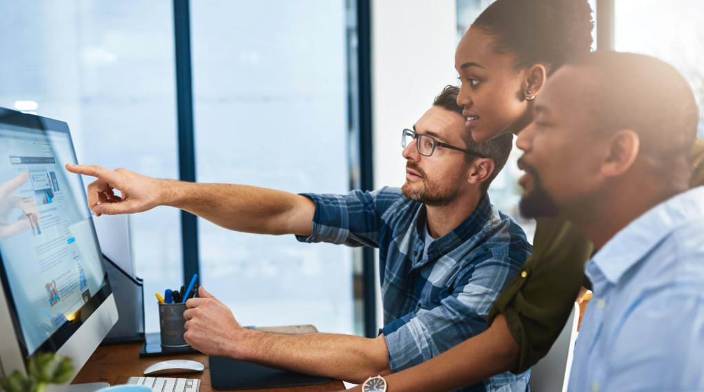 A group of adults is seated in an office around a laptop. One of them points at the screen as the rest of them look.