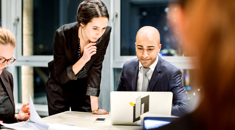 A group of business men and women working in a meeting room. One of the woman leans over to see their computer screen