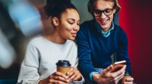 Smiling adult man holding a cellphone and a cup of coffee next to a smiling adult women who is also holding a cup of coffee