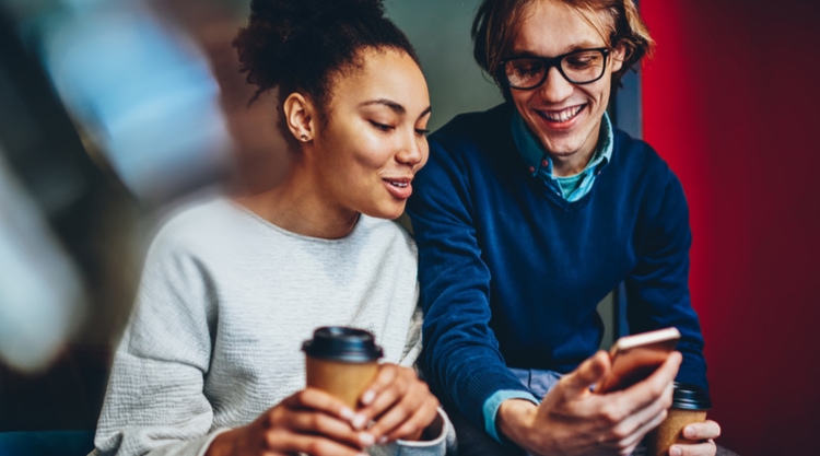 Smiling adult man holding a cellphone and a cup of coffee next to a smiling adult women who is also holding a cup of coffee
