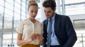 Adult woman typing on her cellphone and adult man looking over while standing in a building