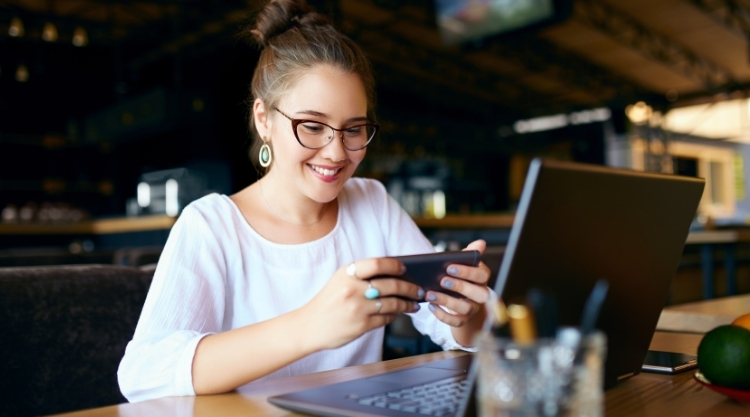 A smiling woman looking down at her cell phone and working on her laptop in a restaurant