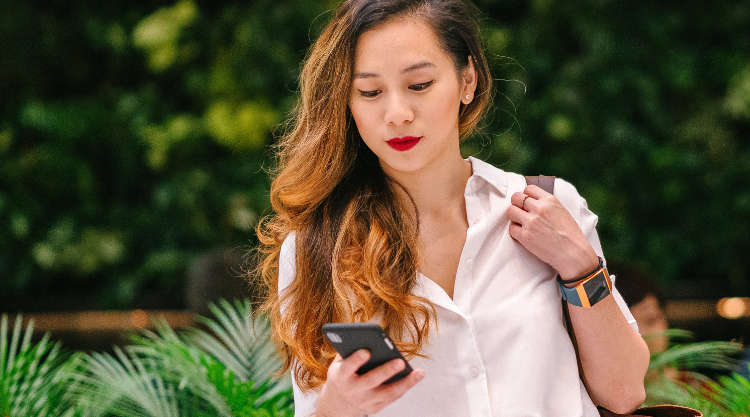 A young woman holding her bag outside of a garden looks down at her phone