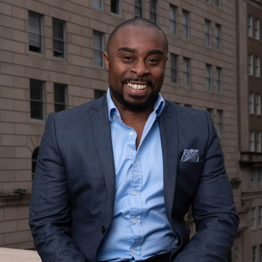 A headshot of a smiling adult man sitting on a ledge. There's a backdrop of a city behind him