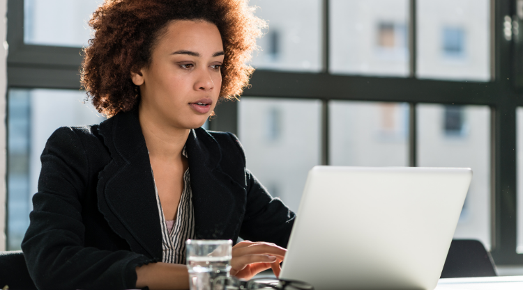 A woman is seated in an office working on her laptop and a glass of water next to her.