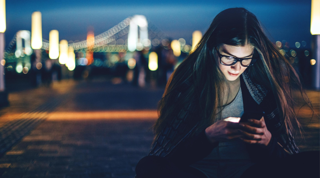 A woman on a dark night outside a city looks down at her phone.