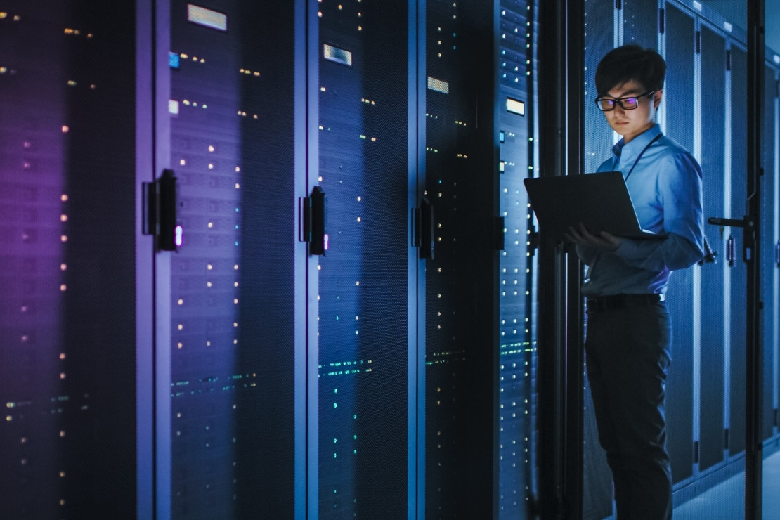 Adult man holding a laptop and standing in a dark room filled with hard drives.