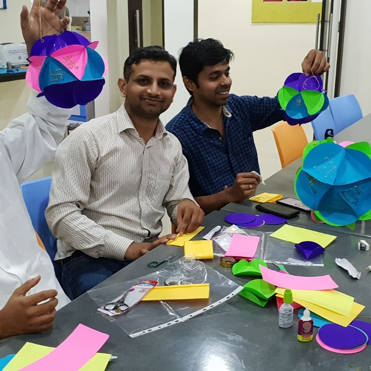 Two adult men sitting at a table. There are arts and craft supplies in front of them. There is the arm of another man holding a completed design