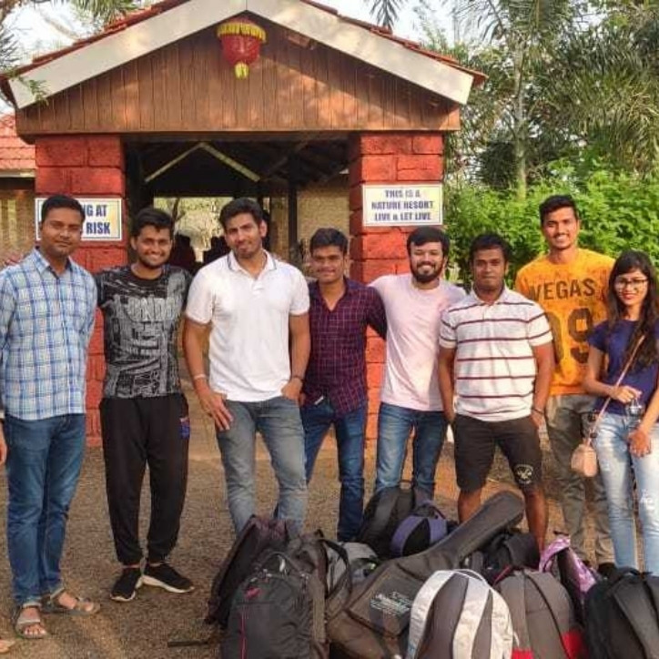 Eight men and a woman standing outside a barn with several book bags and traveling bag in front of them.