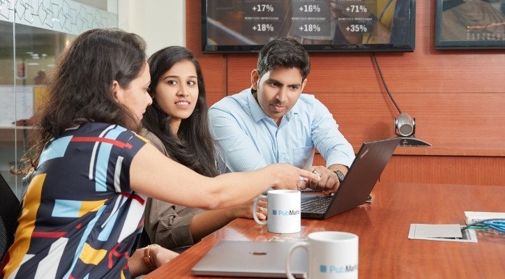 A woman seated by a man and woman, leans towards her laptop and points at her screen. They are in an conference room
