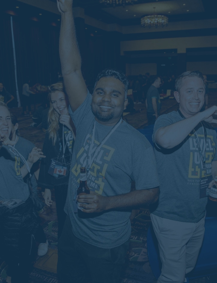 A smiling adult man holding a beer and raising his arm. Next to him is a smiling adult man and two other laughing adult woman as well. There is a dark blue tint over the image