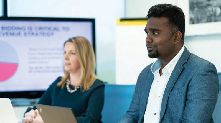 An adult man seated during a presentation next to an older woman speaking