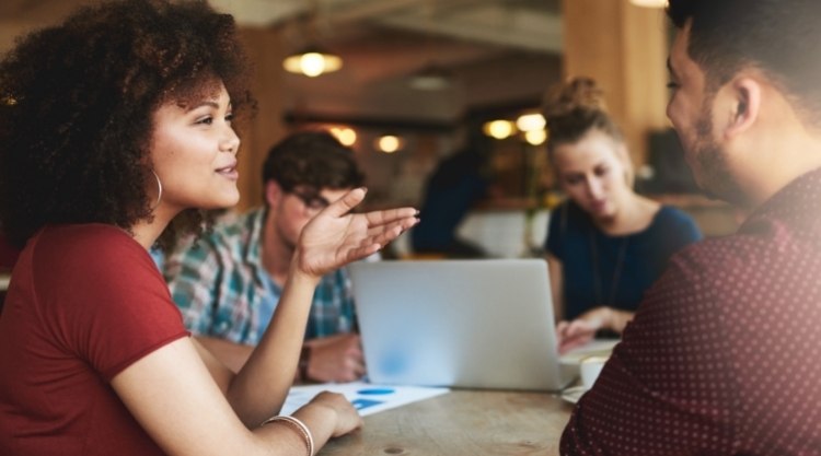 Two people working on a laptop in the background, while two multicultural co-workers discuss work.