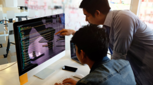 An adult man typing on his computer with another adult man looking over his shoulder and pointing at the screen