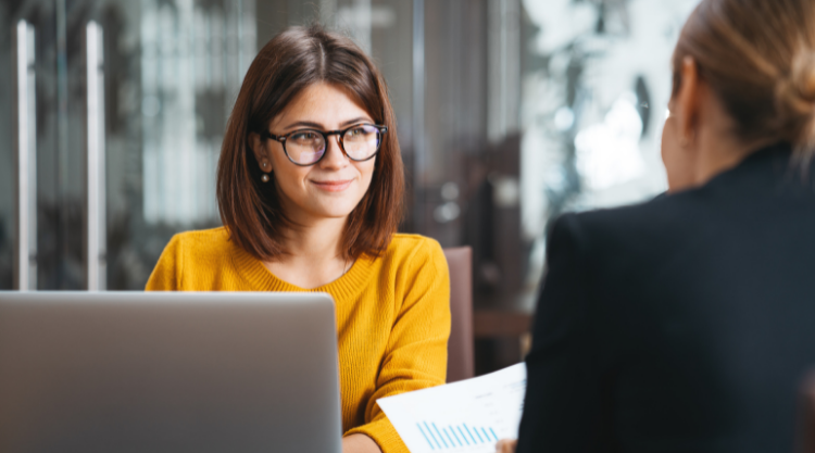 Woman in yellow sweater on laptop discussing work with female co-worker