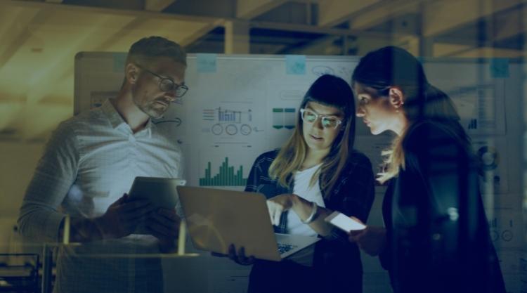 An adult man is holding a tablet and looking at the two women next to him. One woman is holding a laptop, and the other is holding a cell phone and standing in an office space.
