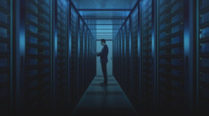 Hall lined with computer hard drives and a tall adult man standing in the center holding a laptop