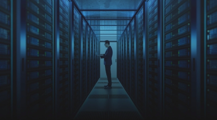 Hall lined with computer hard drives and a tall adult man standing in the center holding a laptop