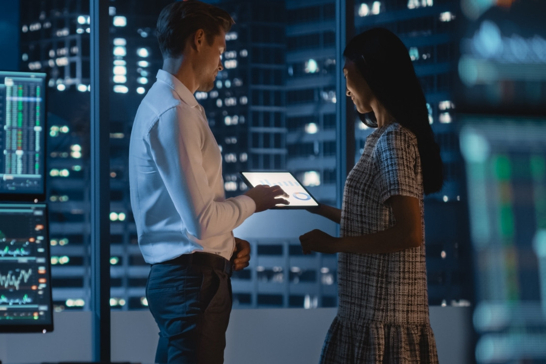 Adult woman looking at te screen the adult man is holding. They are in an office at night in a city