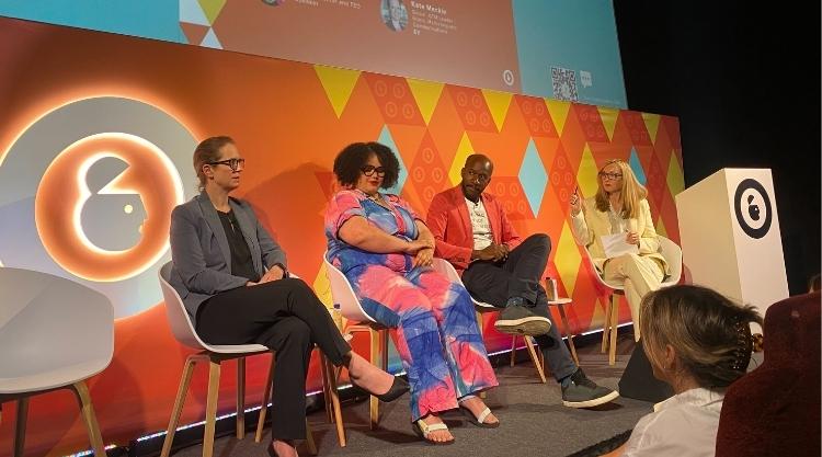 Three women in businesswear sitting on stage during a conference. One man on sitting on stage leans forward.