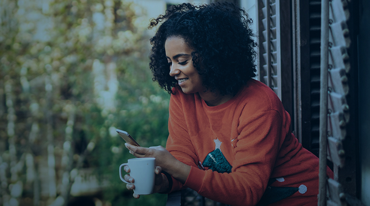 Smiling adult woman looking at cell phone in one hand and holding a mug in the other