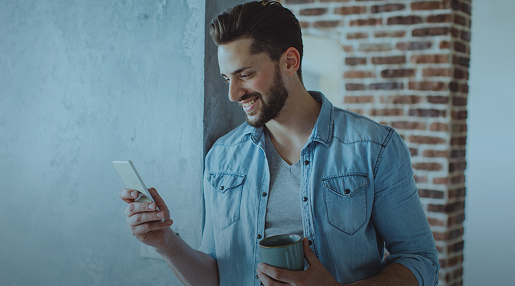 A smiling man using his cell phone and holding a coffee mug while leaning on a wall
