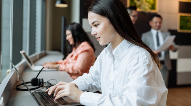 In the foreground, a young woman uses her laptop computer. A colleague of hers works in the background.